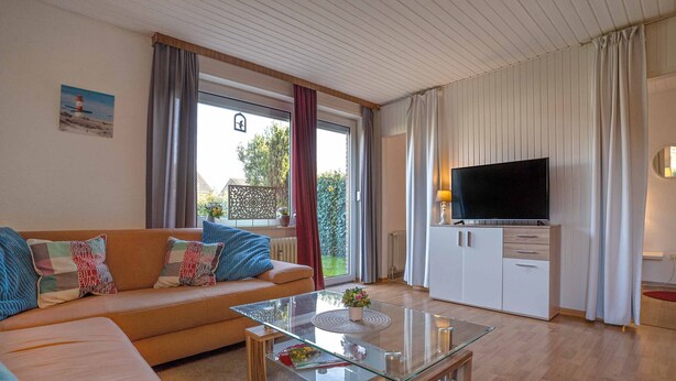 A modern living room with a glass coffee table, a sofa with colorful cushions, a flat-screen TV, and a view of the outdoors.