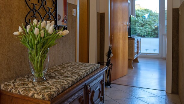 A hallway with a patterned carpet, a wooden bench, a vase with white flowers, and a metal decorative wall piece.