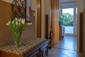 Un pasillo con alfombra estampada, un banco de madera, un jarrón con flores blancas y un adorno metálico en la pared.