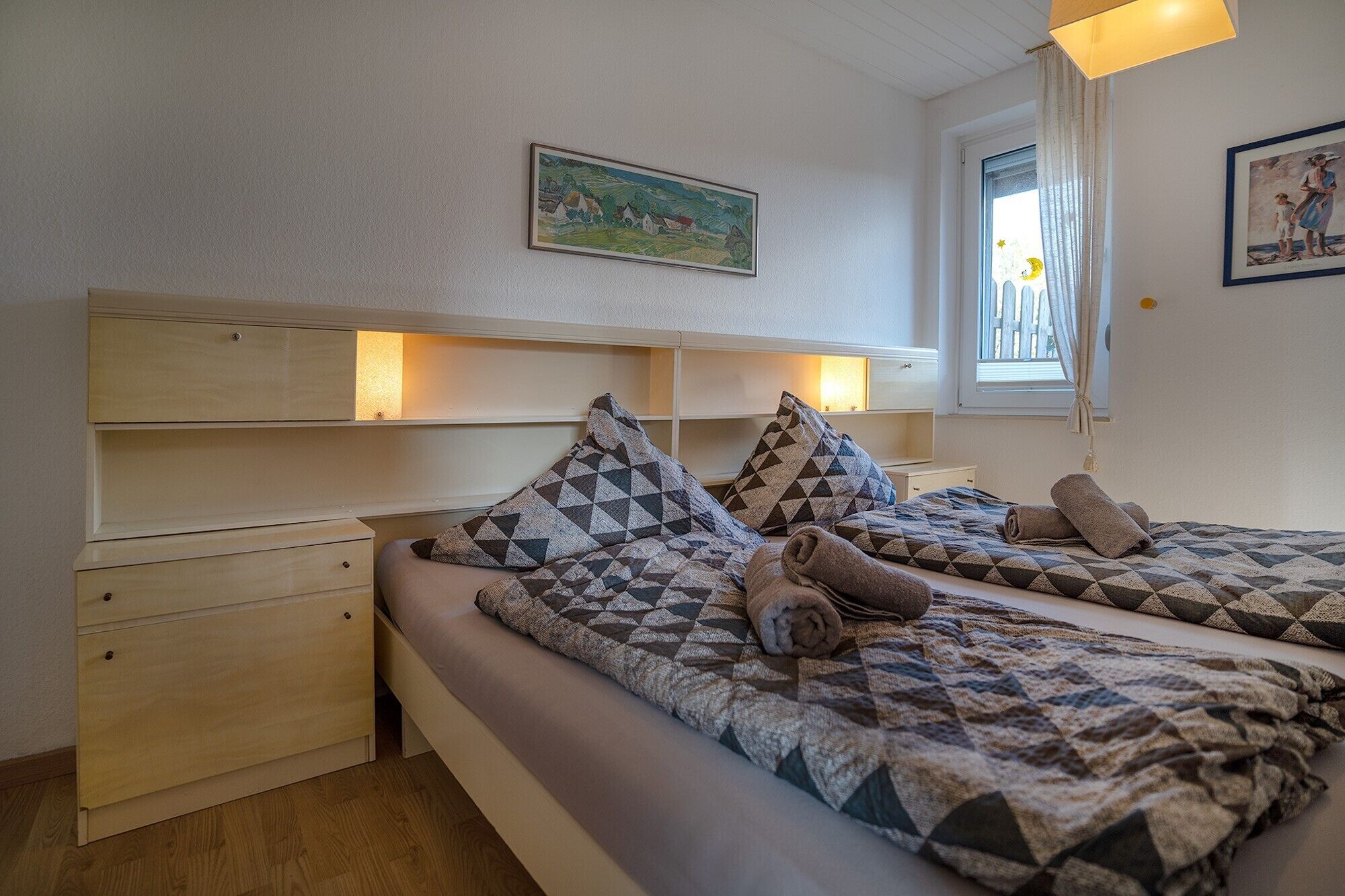 A bedroom with two beds, wooden headboards, a window with curtains, and a painting on the wall.