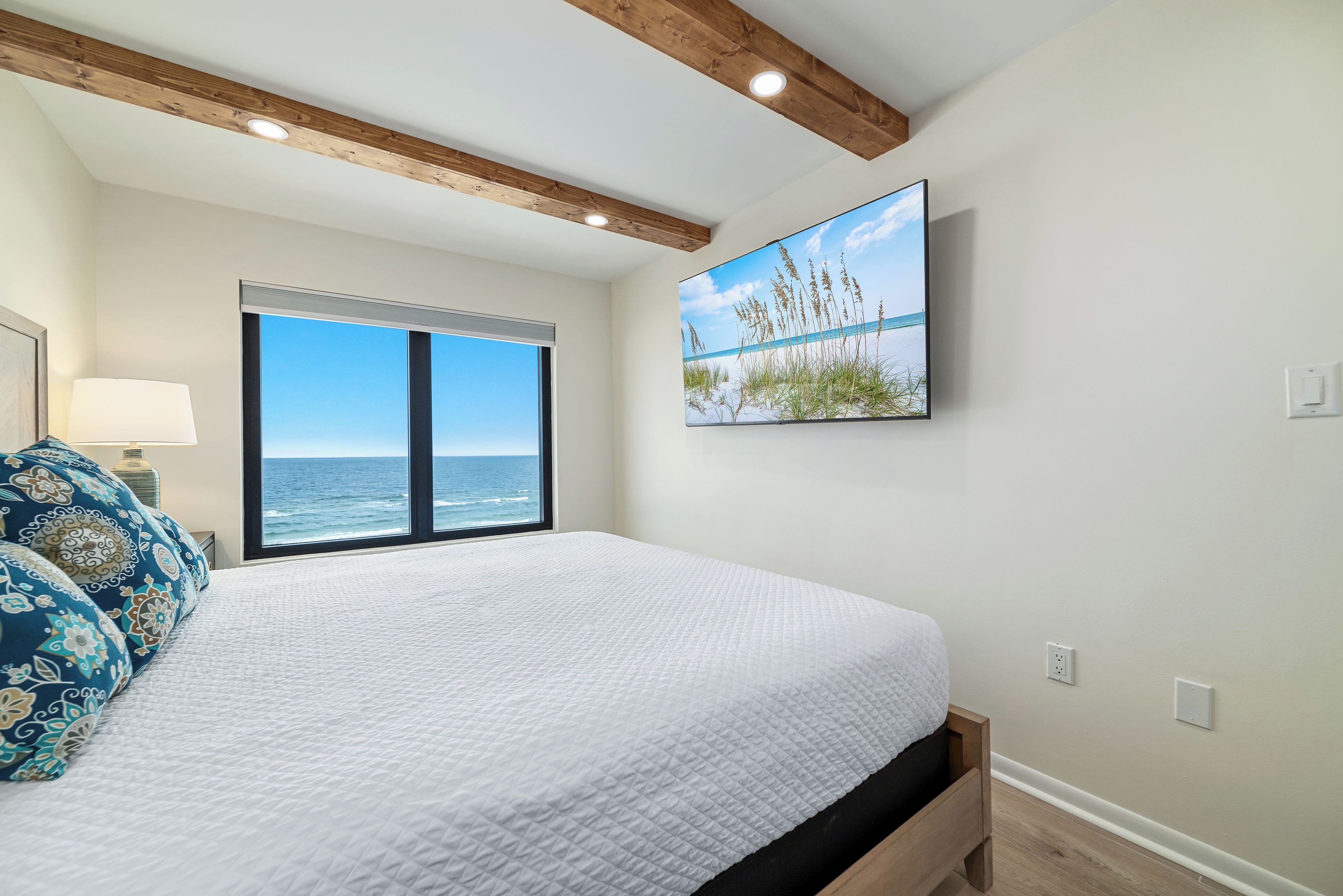 A bedroom with a large bed, a window with a view of the ocean, a painting of grass and water, and wooden ceiling beams.