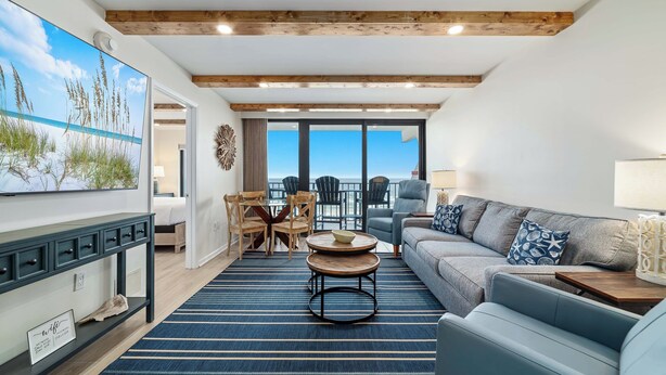 A spacious living area with a large window overlooking the sea, a cozy seating arrangement, and a wooden ceiling. - Island Winds East 608 | Walk to the HangOut! | Beachfront | Pool | My Beach Getaways (Gulf Shores)