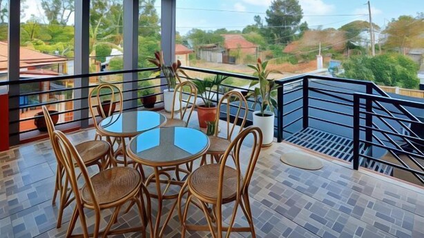 Varanda com mesa de vidro e cadeiras de vime, rodeada por plantas em vasos.