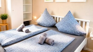 Een slaapkamer met een bed, kussens, een klein tafeltje met een plant en lijstjes aan de muur.