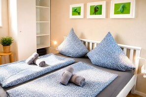 A bedroom with a bed, pillows, a small table with a plant, and framed pictures on the wall.
