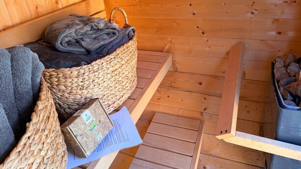 Een houten blokhut vanbinnen met een rieten mand met handdoeken, een houten bank en een klein tafeltje met een boek.