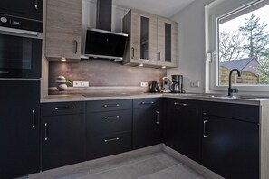 A modern kitchen with dark cabinets, a built-in oven, a microwave, and a window with a view of trees. - Holiday Home 'Schwalbe' with Private Terrace, Garden, and Wi-Fi (Berumbur)
