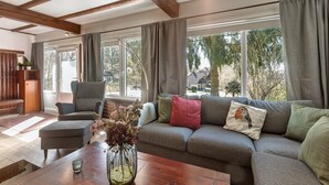 A living room with a sofa, armchair, and coffee table, large windows with curtains, and a wooden ceiling.
