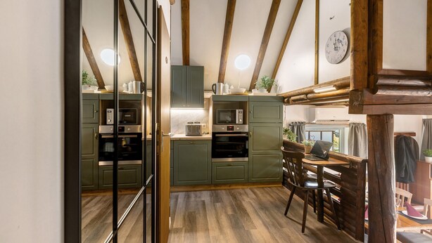 A kitchen with wooden beams, green cabinets, a laptop on a table, and a clock on the wall.