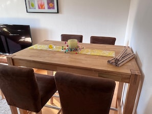 Une table à manger en bois, avec un set de table, un vase en verre contenant des fleurs et un tapis enroulé sur le côté droit.