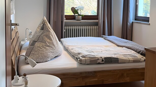 Ein Schlafzimmer mit Bett, Nachttisch, Lampe, Fenster mit Vorhängen, Heizkörper und Blick auf Bäume.