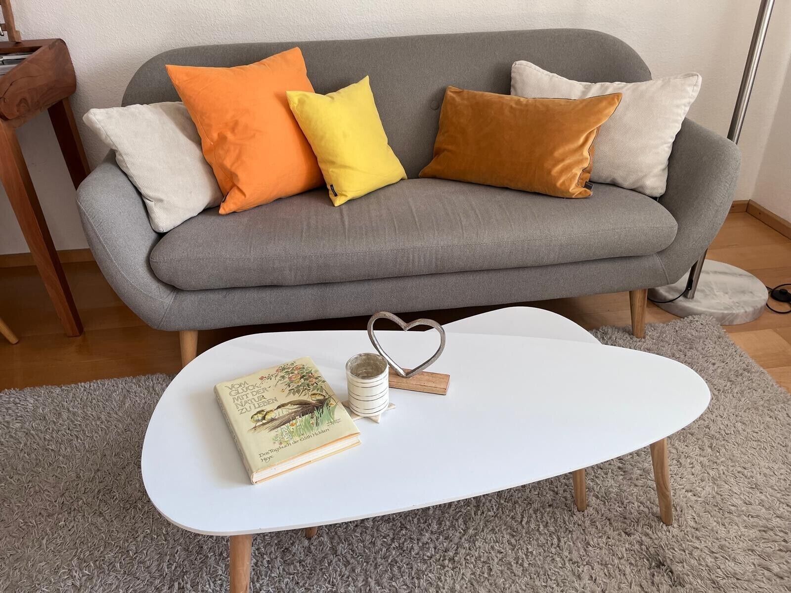 Een grijze bank met oranje, gele en bruine kussens, een witte salontafel met een boek en een hartvormig object, en een grijze vloerkleed.