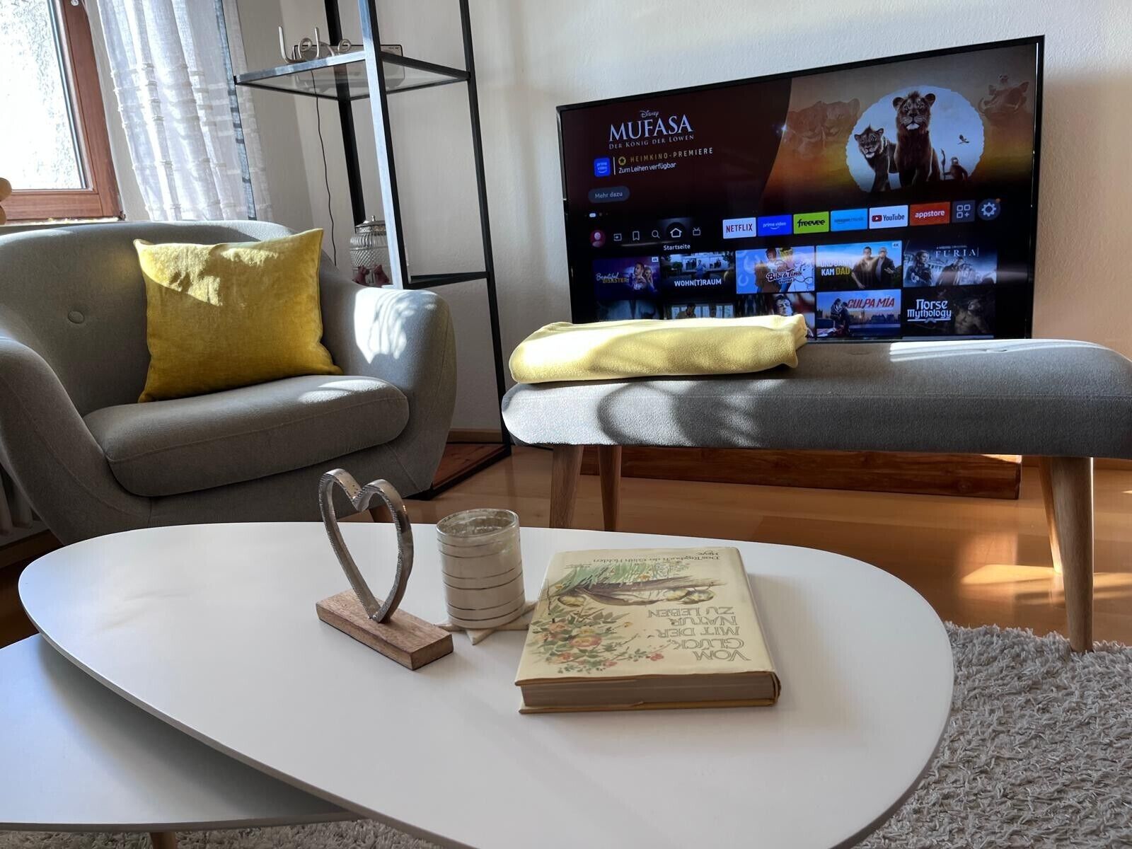 A living room with a flat-screen TV displaying a movie, a grey sofa with yellow cushions, a white coffee table with a book and a decorative metal piece, and a wooden bench.