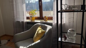 Ein Zimmer mit Fenster, einem Sessel, einem Heizkörper, einem Regal mit Radio und einer Vase mit Blumen.