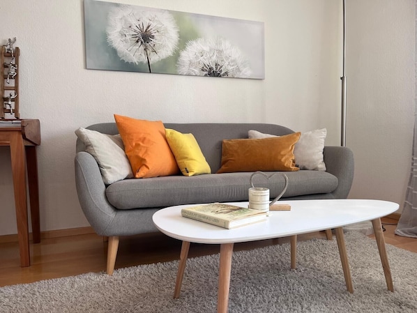 Un salon avec un canapé gris, des coussins colorés, une table basse blanche et un tableau représentant un pissenlit accroché au mur.