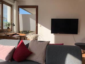 Un salon moderne avec un canapé gris, des coussins rouges et beige, un bureau avec une lampe, une plante en pot et un téléviseur fixé au mur.