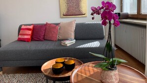 Ein modernes Wohnzimmer mit grauem Sofa, bunten Kissen, einem kleinen Holztisch mit dekorativen Schalen, einer Topfpflanze und einem Gemälde an der Wand.