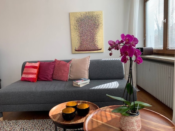 Un salon moderne avec un canapé gris, des coussins colorés, une petite table en bois ornée de bols décoratifs, une plante en pot et un tableau accroché au mur.