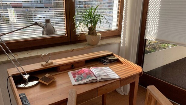 Ein Holztisch mit Lampe, Fernbedienung und Buch, neben einem Fenster mit Jalousien und einer Pflanze.