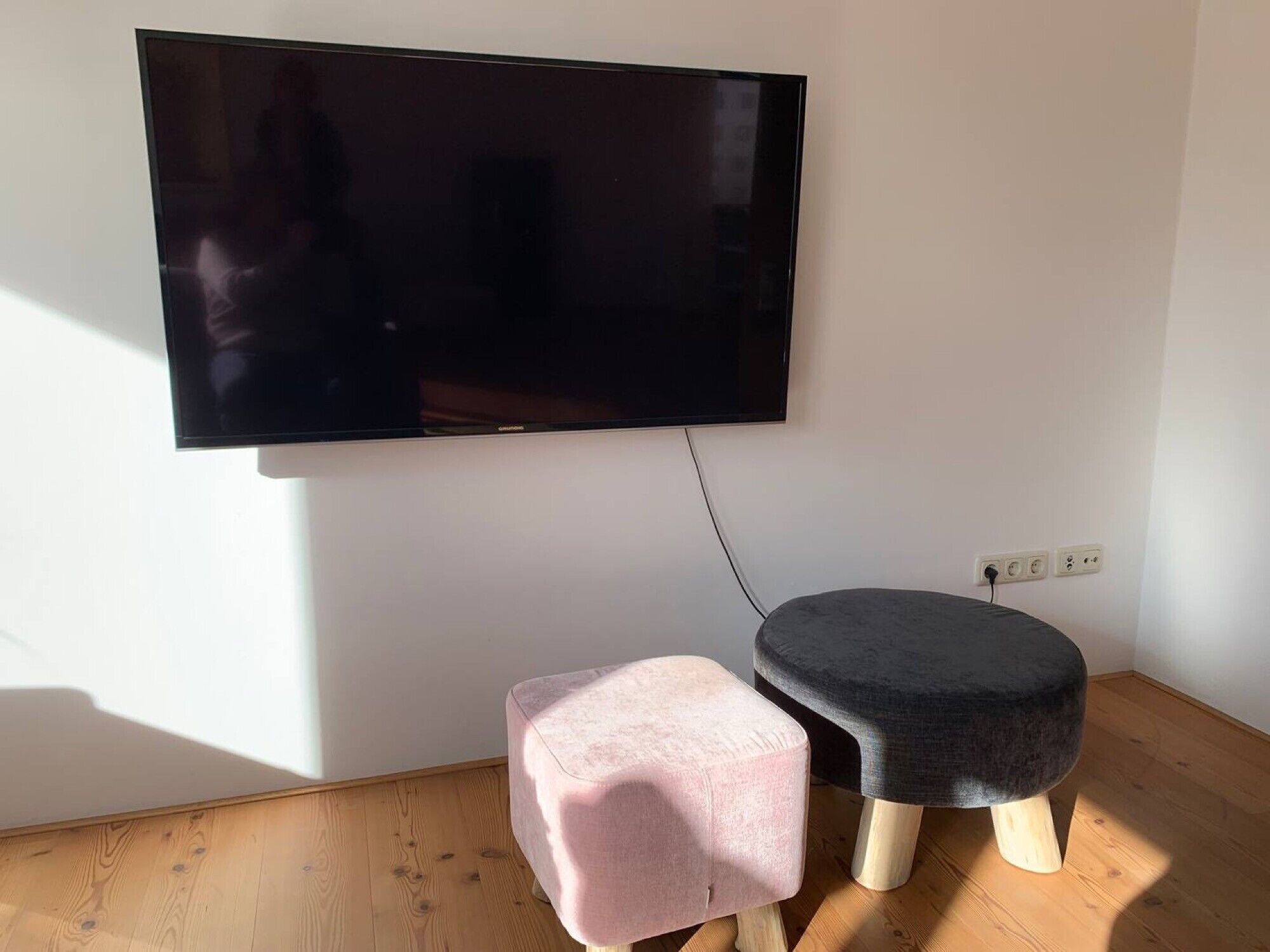A room with a flat-screen TV mounted on the wall, a pink ottoman, and a dark ottoman on the floor.