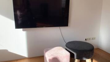 A room with a flat-screen TV mounted on the wall, a pink ottoman, and a dark ottoman on the floor.