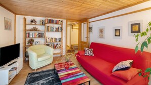 Un salon avec un canapé rouge, un fauteuil de couleur claire, un téléviseur et une bibliothèque remplie de livres et d’objets décoratifs.
