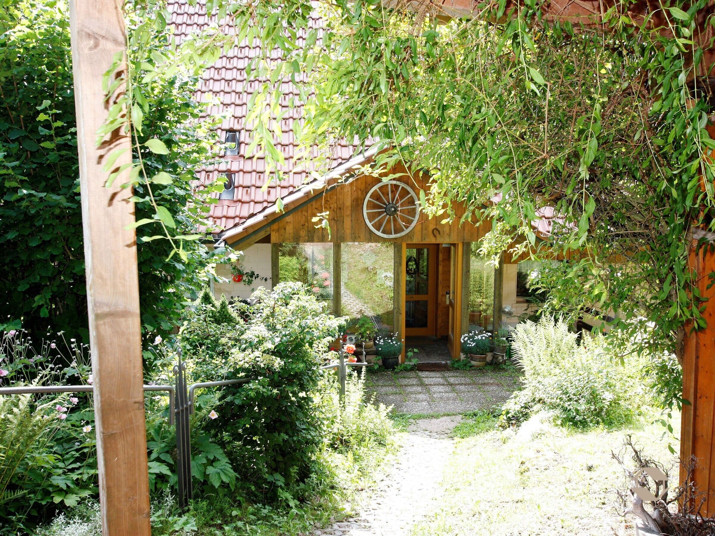 Een houten huis in rustieke stijl met een raam in de vorm van een wiel, omringd door weelderige begroeiing en een grindpad.