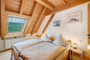 Une chambre avec un plafond en pente, une fenêtre donnant sur un champ verdoyant, un lit avec des oreillers, une table de chevet avec une lampe, et deux tableaux encadrés accrochés au mur.