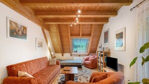 Uma sala de estar aconchegante no estilo loft, com teto de madeira, um sofá, uma poltrona, uma mesinha e uma TV.