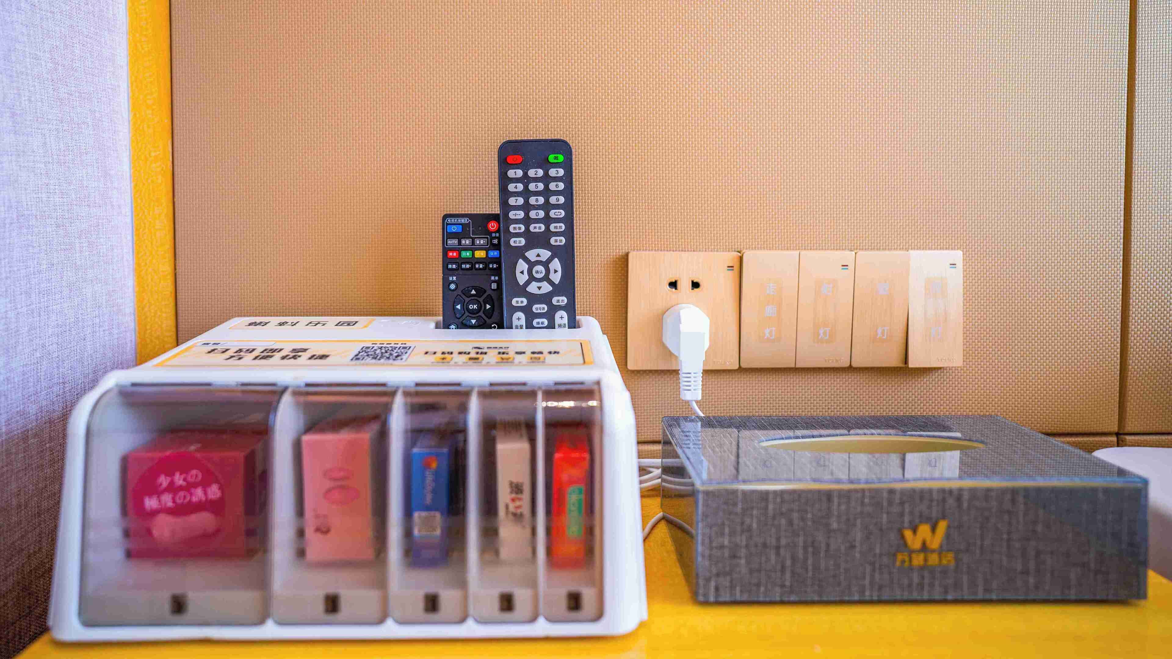A hotel room with a bedside table holding a remote control, a power strip, and a box labeled 'W'.