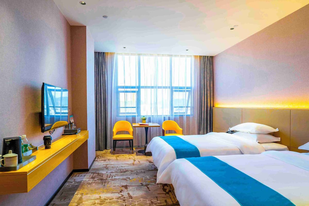 A hotel room with two beds, a desk with a TV, a small table with chairs, and large windows with curtains.