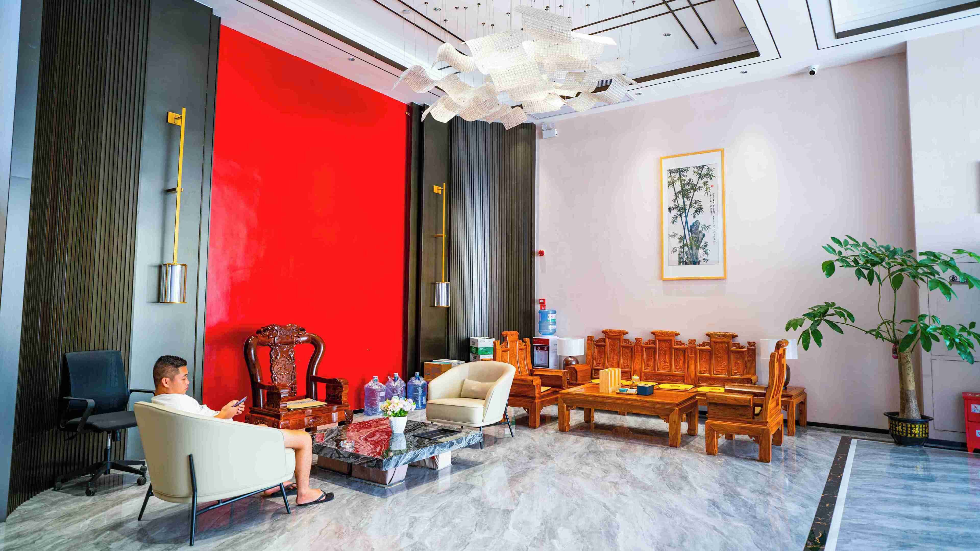 A modern hotel lobby with a red accent wall, a large chandelier, and a traditional wooden sofa set.