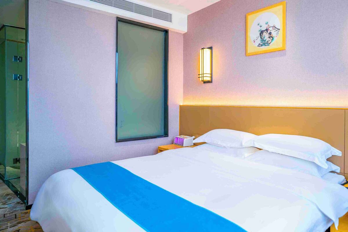 A hotel room with a large bed, a bedside table with a lamp, a wall-mounted artwork, and a window with blinds.