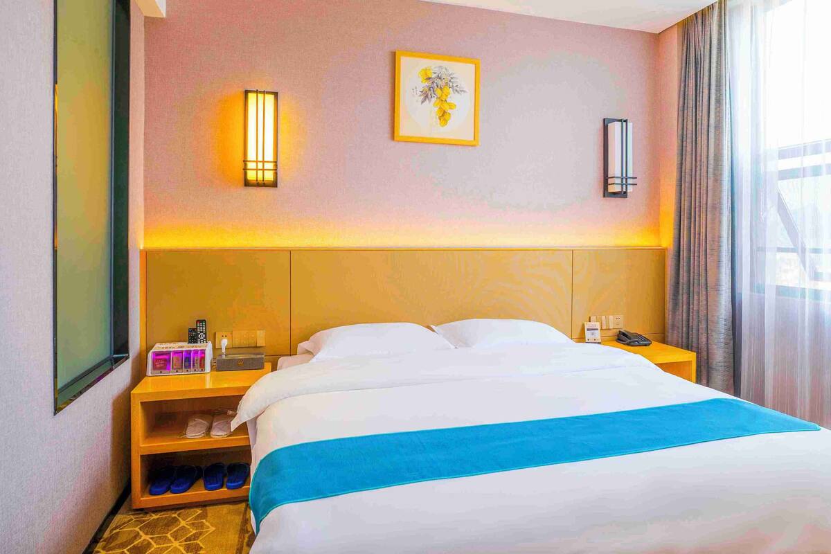A hotel room with a large bed, a bedside table with a phone and a lamp, a small shelf with slippers, and a window with curtains.