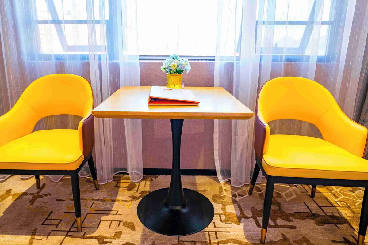 A room with two yellow chairs and a small table with a book and a vase of flowers.