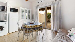 Cozinha compacta com micro-ondas, frigorífico e máquina de lavar roupa. Inclui uma mesa de jantar com cadeiras e um sofá com almofadas. Uma porta de correr envidraçada dá acesso a uma área externa.