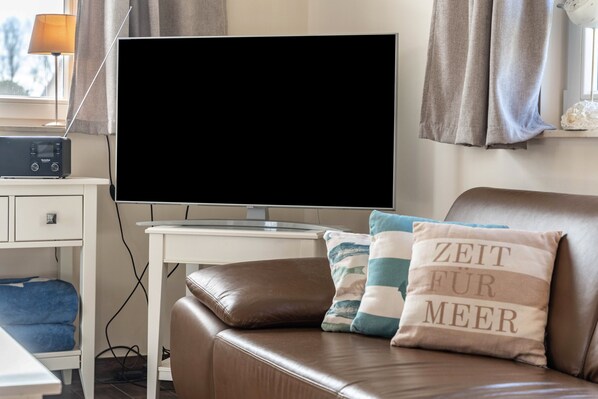 Uma sala de estar com televisão de tela plana, sofá de couro, uma mesinha e um abajur.