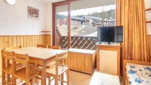 A room with a wooden table and chairs, a television on a wooden stand, and a large window with a view of buildings and hills. - Apartment in France near Ski Resort (Morillon)