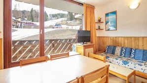A room with a wooden table and chairs, a sofa with patterned cushions, a television on a wooden stand, and a large window with a view of a ski slope. - Apartment in France near Ski Resort (Morillon)