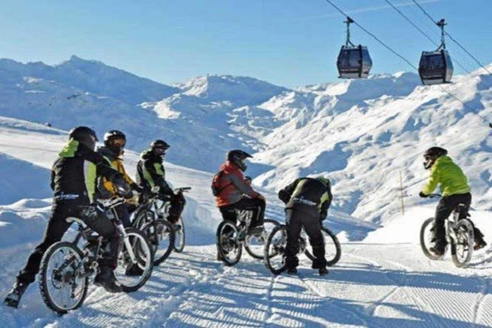A group of people on bicycles on a snowy mountain slope with a ski lift in the background.