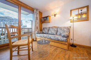 Une pièce chaleureuse avec un plancher en bois, un canapé agrémenté de coussins à motifs, une petite table ronde avec des verres, et un téléviseur posé sur une étagère en bois.
