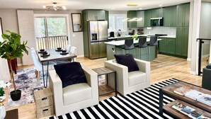 Una cocina moderna con armarios verdes, una isla central y taburetes de bar. Hay un comedor con mesa y sillas, y una sala de estar con sofá y mesa de centro.