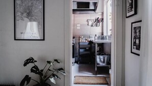 A hallway leading to a kitchen with a potted plant, a framed picture on the wall, and kitchen utensils hanging above the counter.