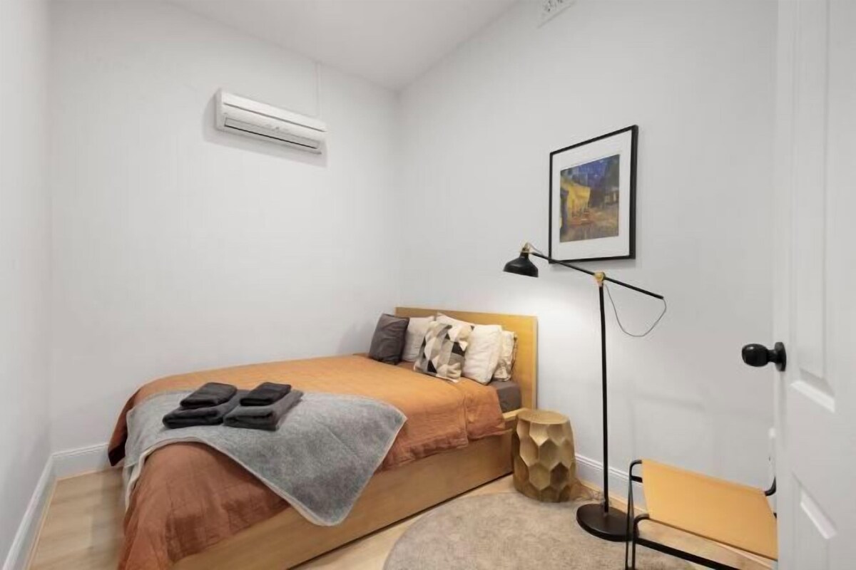 Un dormitorio con cama, aire acondicionado, mesita de noche, una silla y un cuadro enmarcado en la pared.