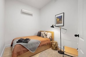 A bedroom with a bed, an air conditioner, a bedside table, a chair, and a framed picture on the wall.