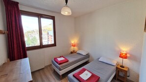 Ein kleiner, hell eingerichteter Raum mit zwei Einzelbetten, einem Holztisch und einem Fenster mit Blick auf Gebäude.