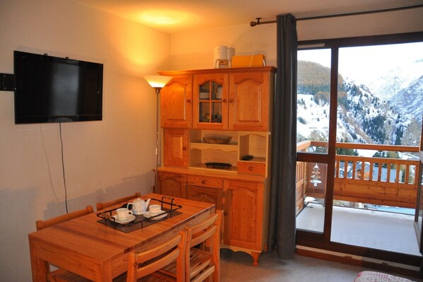 A wooden dining table with a tray, a television mounted on the wall, wooden cabinets, and a balcony with a mountain view.