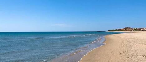 A beach with clear blue water, a sandy shore, and a distant town.