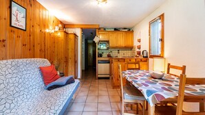 Um quarto aconchegante com parede de madeira, um sofá com capa estampada em folhagens, uma mesa de jantar com toalha de mesa estampada e uma área de cozinha com armários de madeira e micro-ondas.