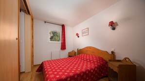 Een slaapkamer met een houten bed, nachtkastje, een rode sprei, een raam met uitzicht op groen en een rood gordijn.